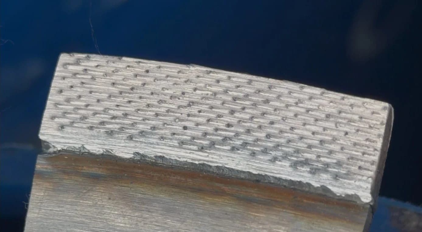 A close-up of a real Exakt segment lying on the brushed steel core of a blade, on a blue ground: dark diamonds set in an even grid across the pale silver segment face, with the weld seam running below it.